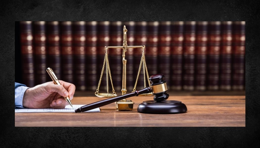 Scales of justice and a gavel on a desk in front of law reports, with a hand writing on a notepad.